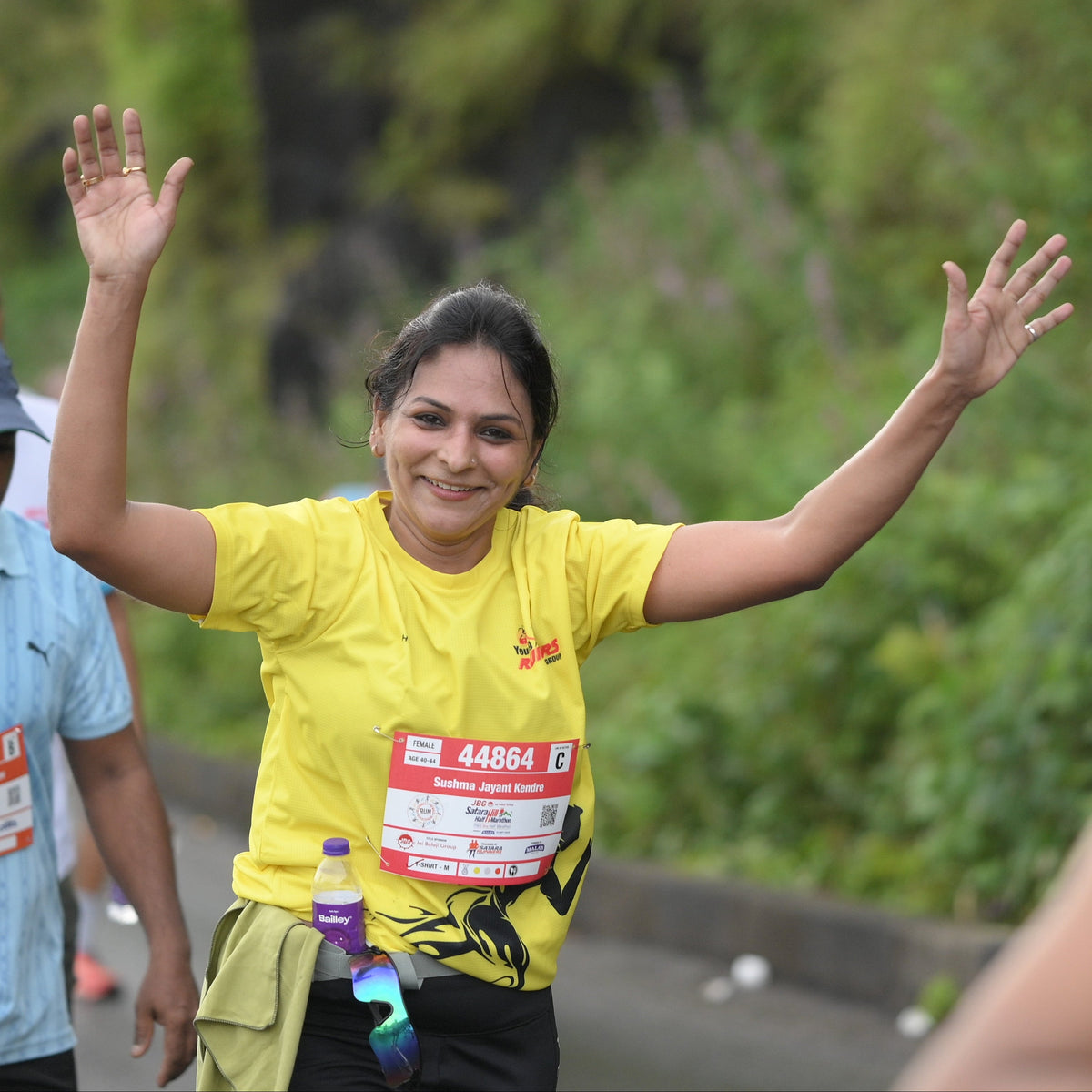 A woman smiling and waving with both arms raised, wearing a bright yellow athletic T-shirt and a race bib outdoors on a path with green foliage in the background.